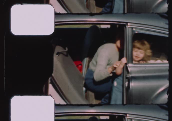 A cute toddler with curly blonde hair stands inside the open door of a vintage car in 1950s home movie footage