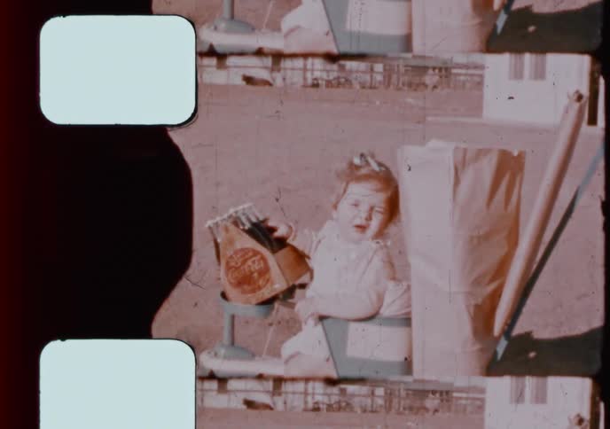 A vintage home movie captures a baby girl sitting in a toy stroller while holding a classic Coca-Cola carton