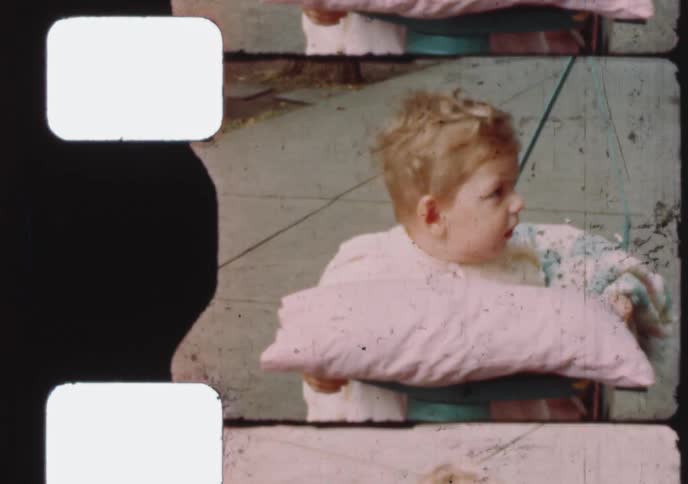 A vintage home movie captures a close-up portrait of an adorable toddler sitting outside in a bouncer
