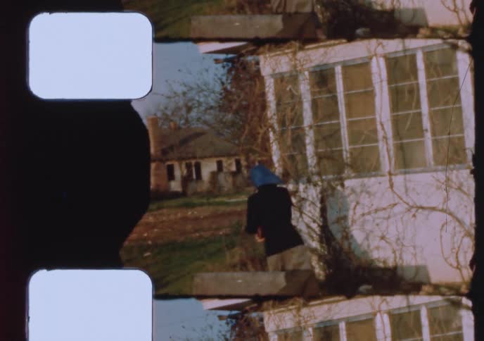A vintage 16mm film shows a young woman in a headscarf smiling as she leaves her home and walks across the yard