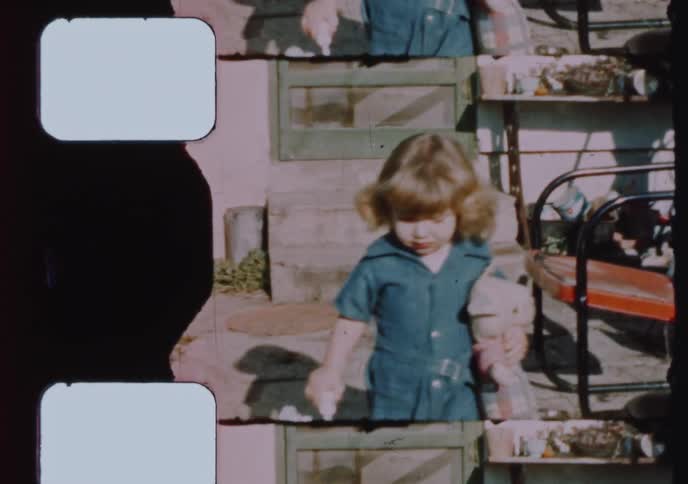 An adorable little girl in a blue jumpsuit holds her stuffed animals and walks toward the camera in a vintage home movie