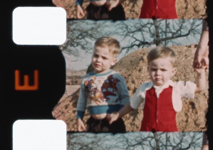 An archival 1950s home movie shows two young brothers holding hands and waving at the camera outdoors