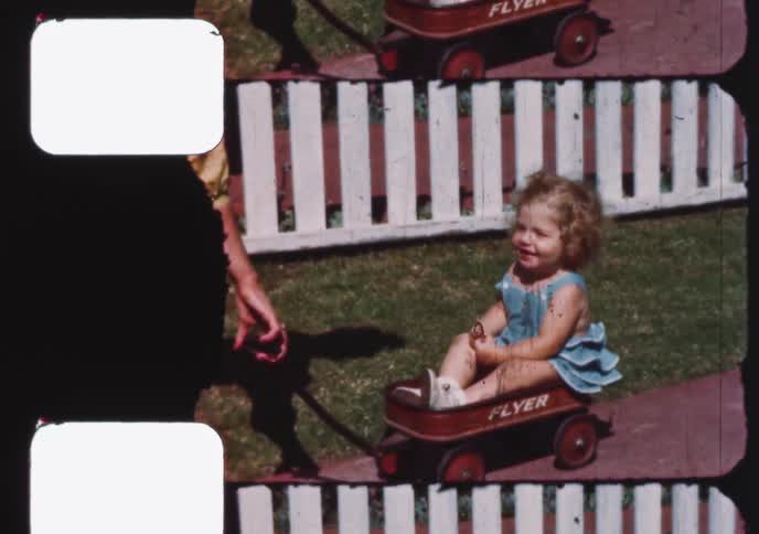 An adorable curly-haired toddler waves and smiles while sitting in a classic red wagon in a sunny backyard