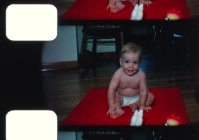 A vintage 8mm home movie from the 1960s captures a mother helping her baby learn to walk, followed by the baby sitting happily on a red mat