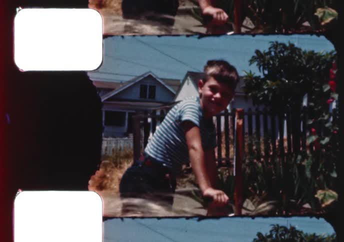 Vintage home movie captures a brother and his toddler sister playing with a tricycle on a sunny suburban sidewalk