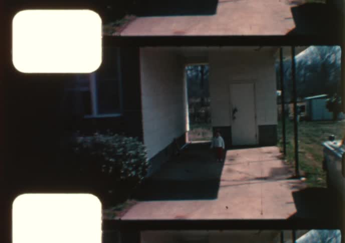 An adorable toddler in a red hat walks towards the camera and then runs away in a driveway in this authentic 8mm home movie
