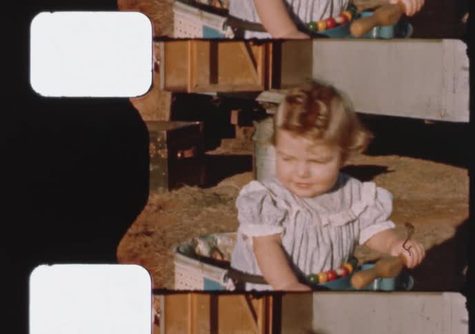 An adorable smiling toddler in a pretty dress plays in her walker outside on a sunny day in this vintage 8mm home movie
