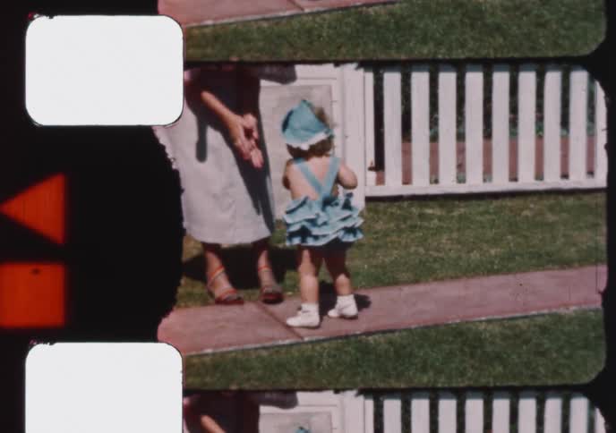 An adorable curly-haired toddler in a frilly blue playsuit and hat explores the backyard on a sunny day in this vintage home movie