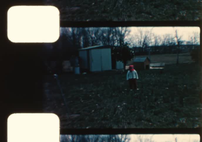 An adorable smiling toddler wearing a red hat and pants walks around the yard in this charming vintage home movie