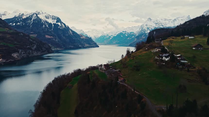 A stunning aerial view slowly pushes in over a vast alpine lake, revealing majestic snow-capped mountains and a serene village on the green hillside