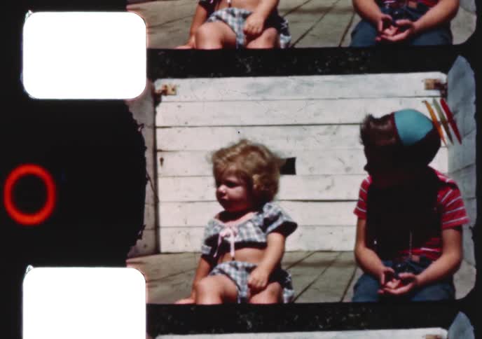 A vintage 8mm film captures a young boy making funny faces in a party hat and a toddler girl playing in their backyard on a sunny day