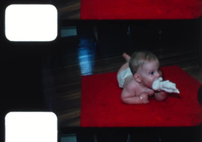 A cute baby in a diaper happily plays with a toy while lying on a red mat in this vintage home movie