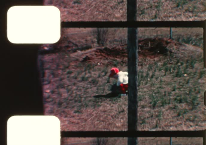 A vintage home movie captures a toddler getting off their bicycle and playing with a ball in the backyard of a suburban home