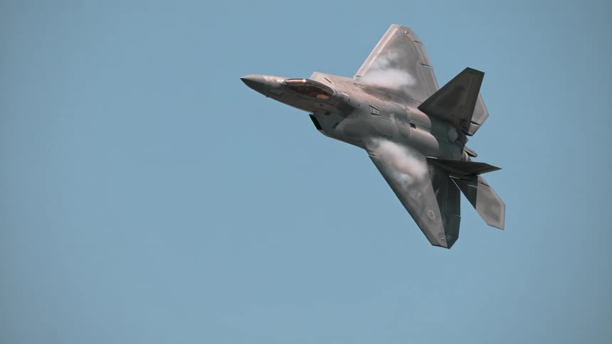An F-22 Raptor fighter jet performs a slow aerial maneuver against a clear blue sky, with vapor forming over the wings as it turns