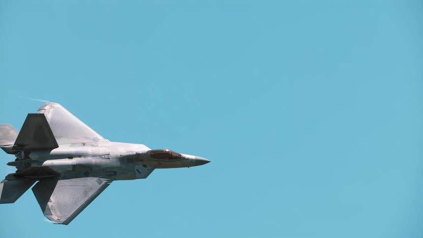 A military F-22 Raptor fighter jet flies in slow motion with afterburners on, creating vapor trails against a clear blue sky
