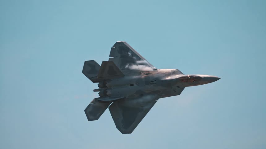An F-22 Raptor fighter jet flies in slow motion with afterburners engaged against a clear blue sky