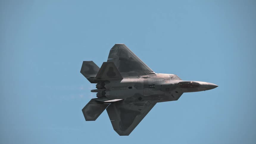 A sleek F-22 Raptor fighter jet soars through a clear blue sky in slow motion, with afterburners glowing and vapor forming over its wings