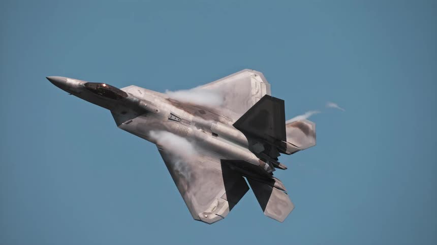 A slow-motion close-up of an F-22 Raptor fighter jet performing a high-G turn, creating vapor trails over its wings and firing afterburners