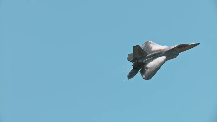A sleek F-22 Raptor performs a high-G maneuver in slow motion, creating impressive vapor trails against a clear blue sky