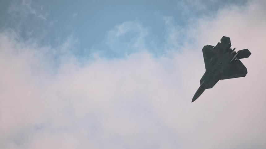 Static low-angle shot of an F-22 Raptor fighter jet suspended in the sky as clouds drift past