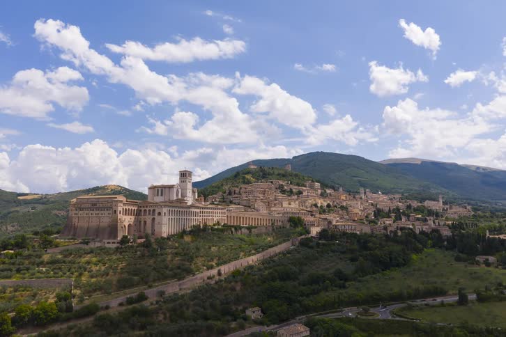 An aerial shot pans up over the historic city of Assisi, Italy, nestled in the green hills of Umbria under a partly cloudy sky