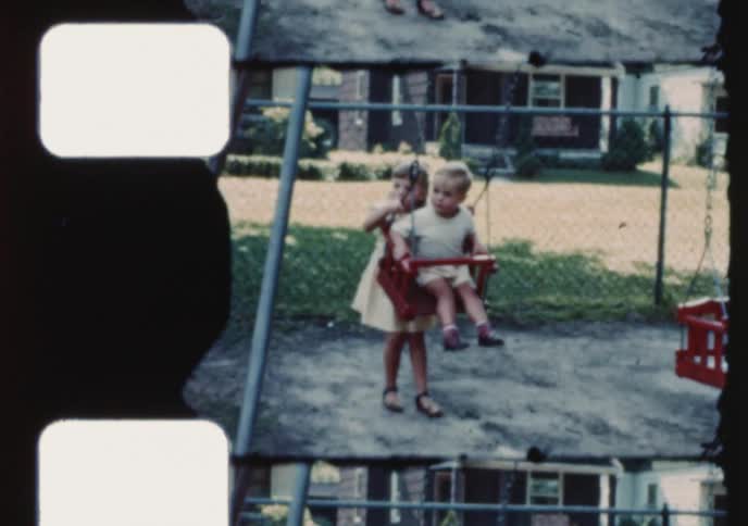 Vintage home movie captures a young girl pushing her little brother on a swing set in a suburban backyard