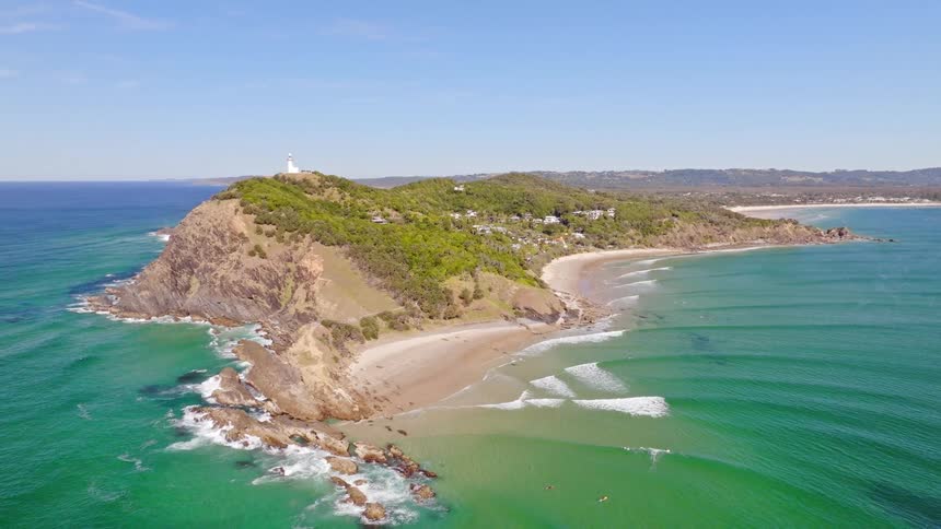 An aerial pullback reveals the iconic Cape Byron lighthouse on a verdant headland surrounded by turquoise ocean waves