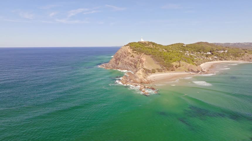 A soaring aerial view of the turquoise ocean as it approaches a beautiful coastal headland with a lighthouse