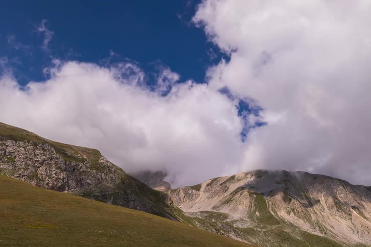 A majestic time-lapse captures billowing white clouds moving across a deep blue sky over a rugged mountain landscape