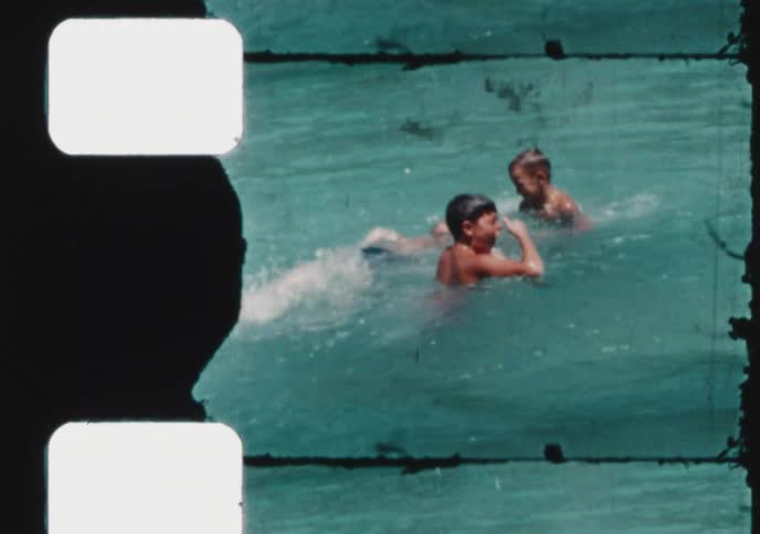 A vintage home movie shot shows children playfully splashing and swimming in turquoise water on a sunny day