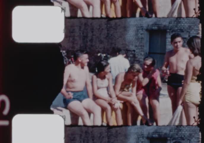 A group of young people in swimsuits laugh and socialize while sitting on a wall in the summer sun, captured on vintage film