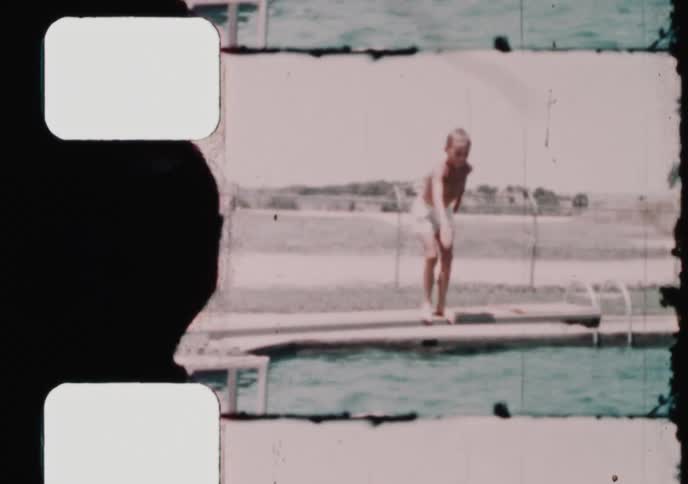 A young boy jumps off the diving board into the swimming pool where his father is waiting on a sunny summer day in this vintage 8mm home movie