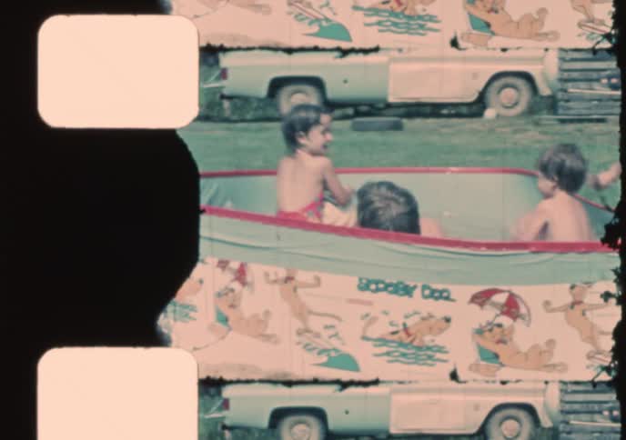 A vintage 8mm home movie shows several happy toddlers splashing and playing in a Scooby-Doo wading pool in their backyard