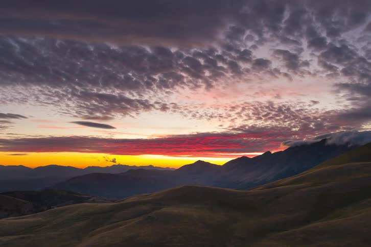 A dramatic sunset paints the textured clouds in fiery red and orange hues above a serene landscape of rolling hills and distant mountains