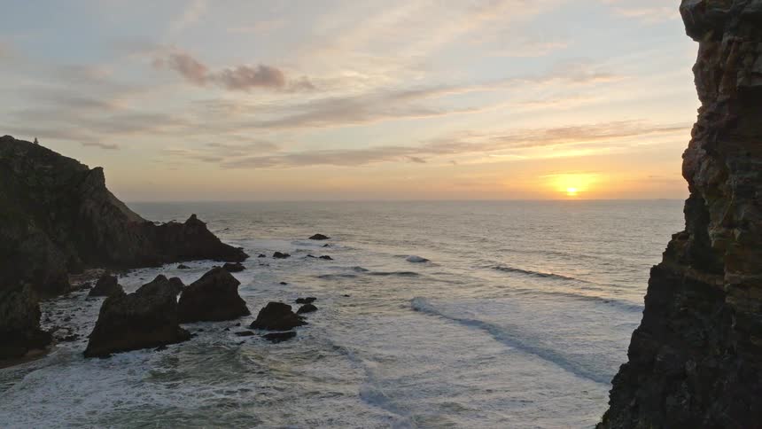 An aerial shot rises from behind a large rock formation to reveal a beautiful sunset over a rugged, wavy coastline