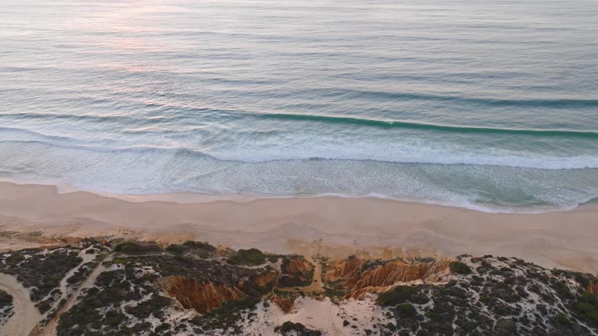 A tranquil aerial view tilts up from a sandy beach and cliffs to reveal the vast ocean during a beautiful sunrise