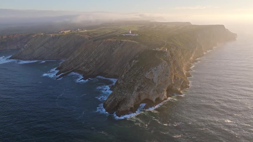 A static aerial view shows a lighthouse perched on a dramatic, rugged cliffside peninsula during a hazy golden hour