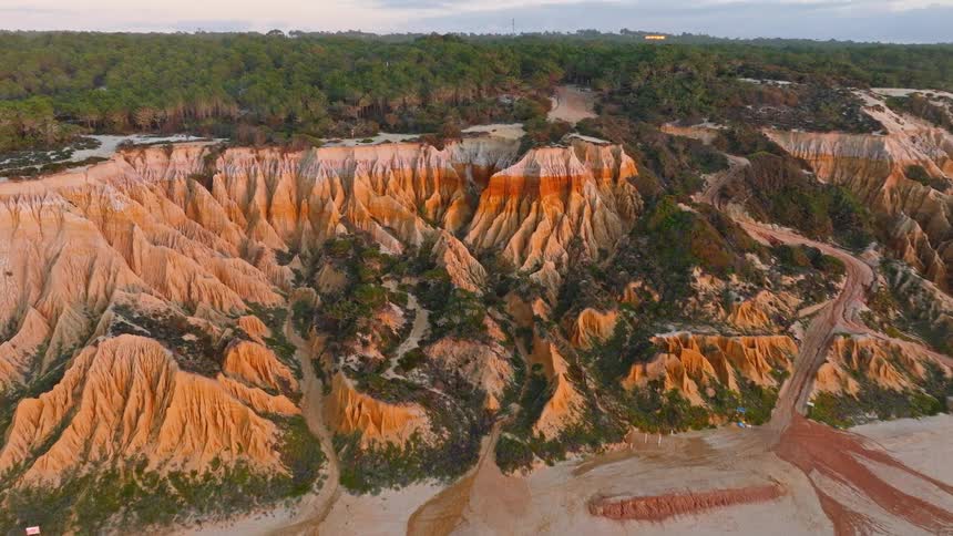 A stunning aerial view of uniquely eroded multicolored cliffs meeting a sandy beach and lush forest at golden hour