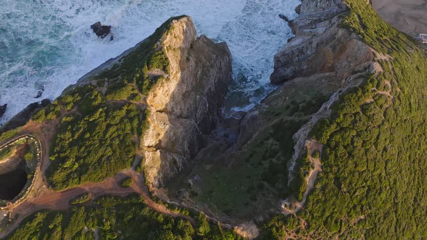 A stunning aerial view captures two lush green headlands separating the powerful crashing waves of the ocean from a tranquil inlet at golden hour