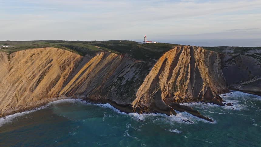 An aerial pullback shot reveals a majestic lighthouse standing atop dramatic stratified coastal cliffs at golden hour