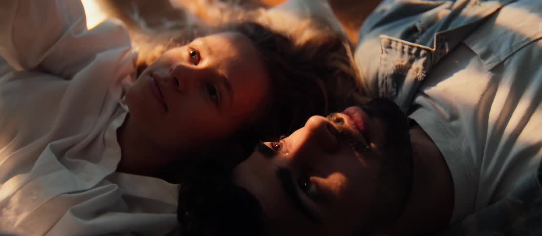An intimate high-angle shot of a young couple lying together, their faces illuminated by the warm, golden light of the sun