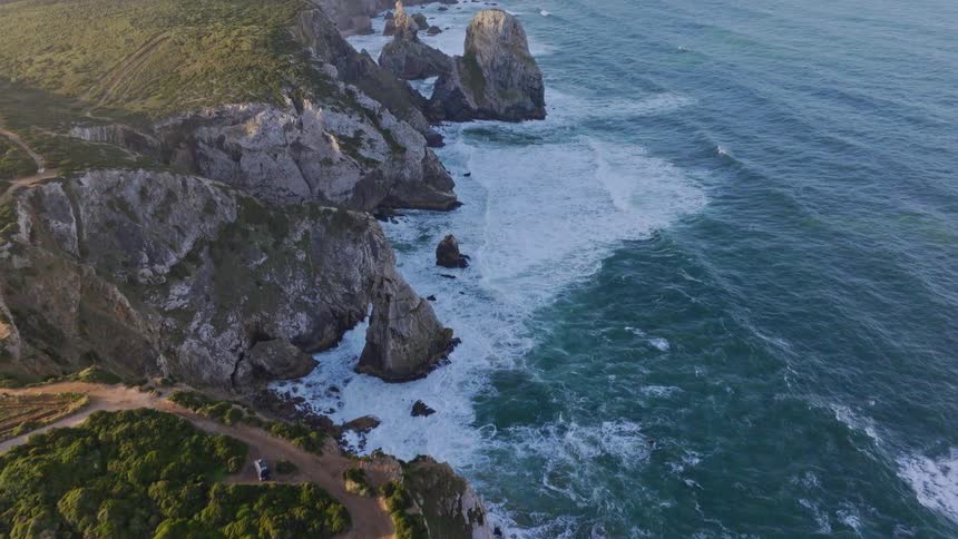 An epic aerial reveal of a rugged, green coastline with dramatic sea stacks, crashing waves, and a distant lighthouse at golden hour