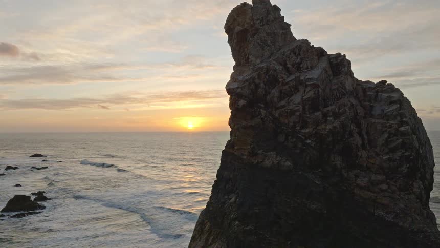 A majestic rock formation stands silhouetted against a beautiful ocean sunset as the camera slowly pulls back