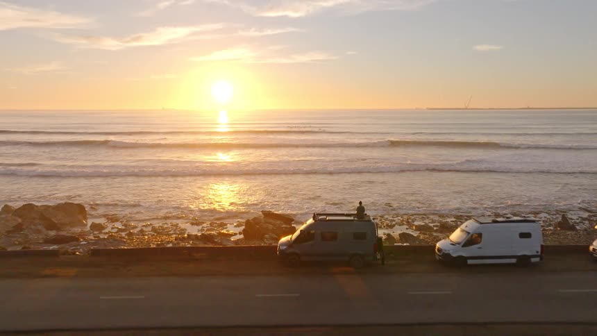 An aerial view flies past camper vans parked on a coastal road to reveal a stunning golden sunset over the ocean