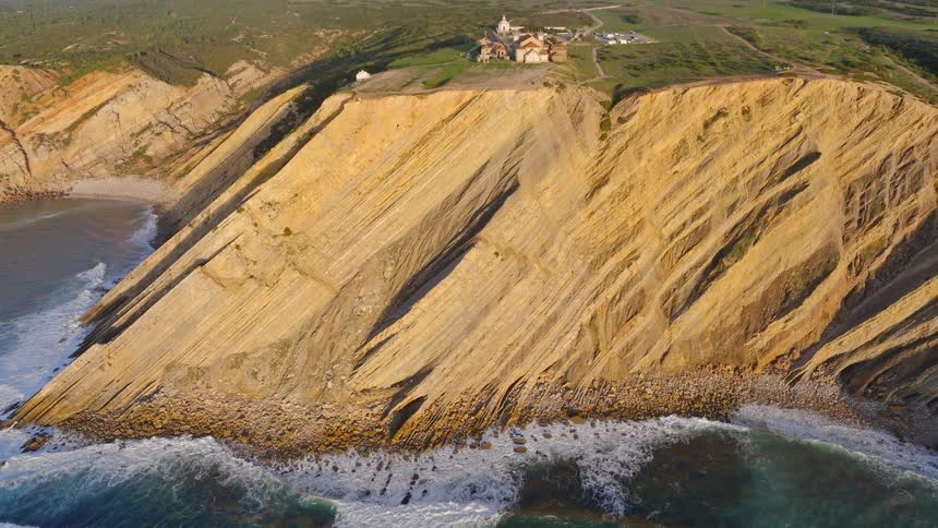 An aerial view pulls back from dramatic, layered cliffs to reveal a historic sanctuary perched on the coastline as waves crash below