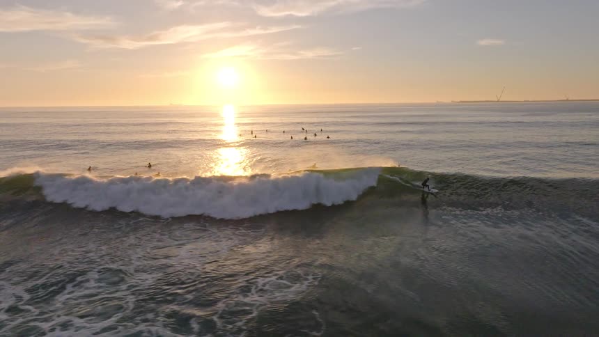 In a stunning slow-motion aerial shot, a surfer catches a perfect wave and rides it towards the golden sunset