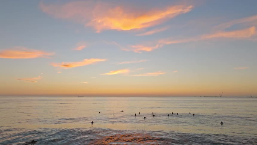A serene view shows a group of surfers floating in the calm ocean, waiting for waves under a beautiful sunset sky