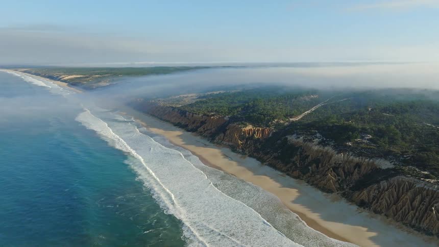 A stunning aerial view captures a vast coastline with turquoise waves, sandy beaches, and a thick layer of fog rolling over the forested cliffs