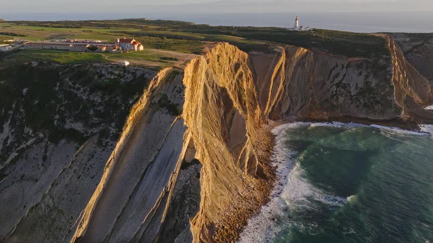 A breathtaking aerial shot rises to reveal the majestic cliffs and lighthouse of a scenic coastline during a beautiful sunset