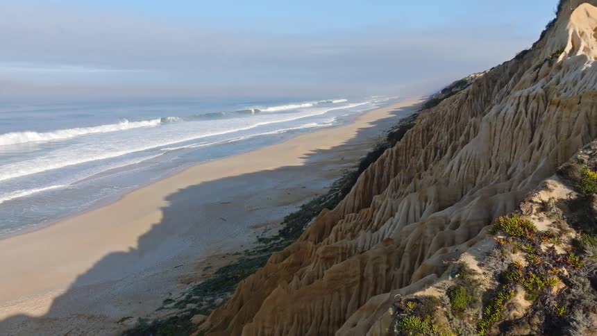 Aerial footage slowly reveals the stunning, eroded sandstone cliffs of a deserted beach on a sunny, hazy day